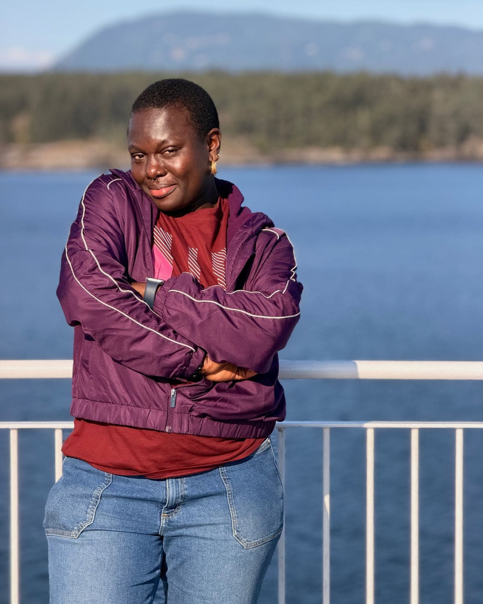 Her on a ferry rail in a plum windbreaker, arms folded, blue water and islands behind