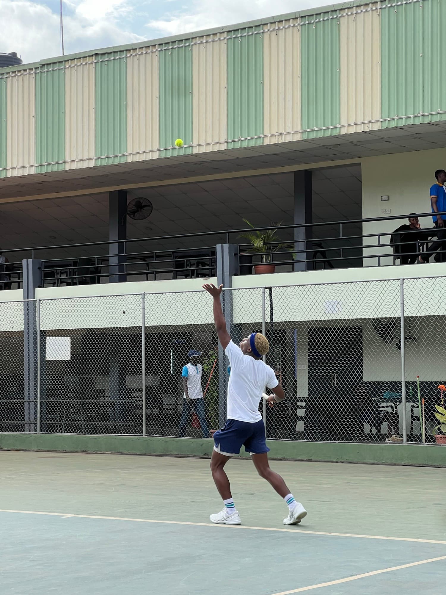 Serving on an outdoor club court, ball floating above the clubhouse rail
