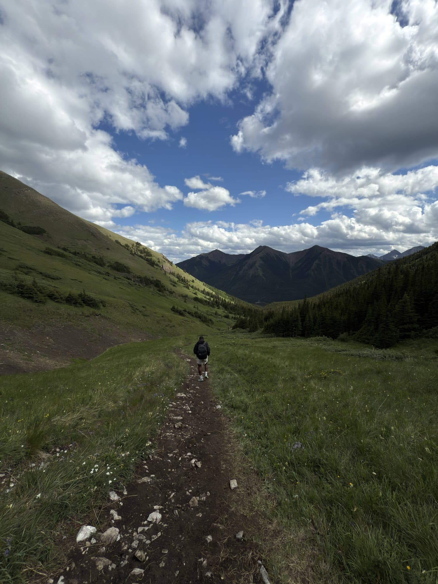 A lone hiker on a dirt trail winding toward distant peaks under a big cloudy sky