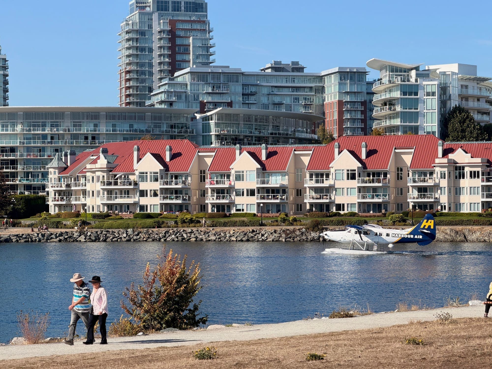 A seaplane coming in low over waterfront townhouses as two people watch from the shore