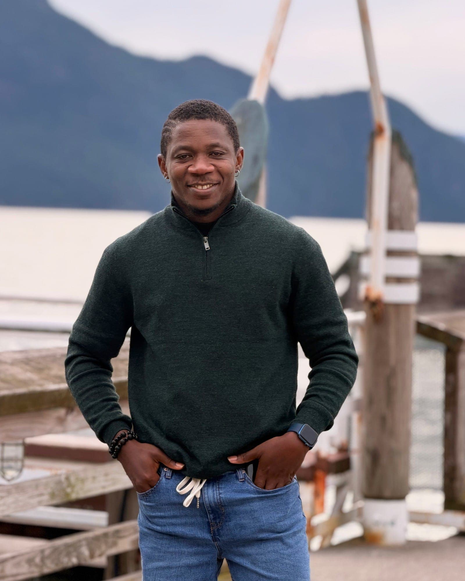 Standing on a pier in a green quarter-zip, hands in pockets, mountains soft behind