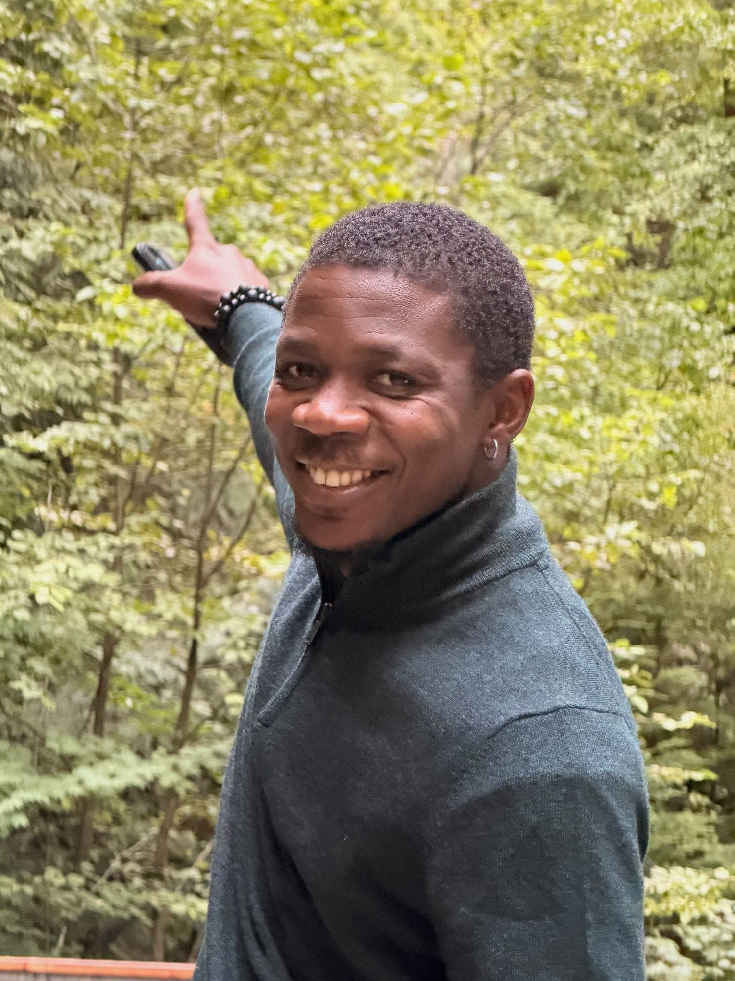 Grinning over one shoulder and pointing back at a wall of green trees