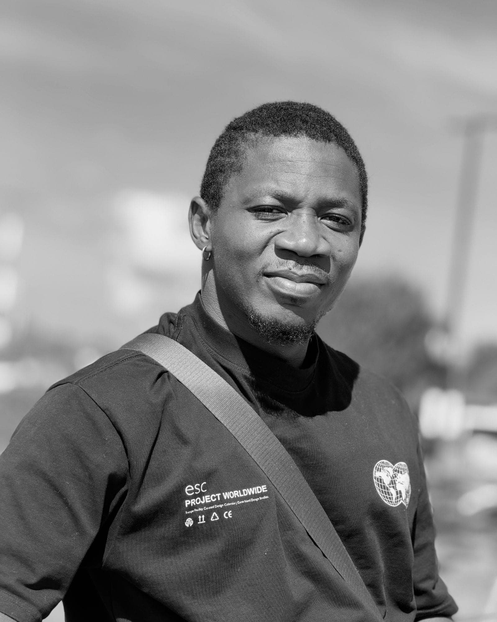 A black and white portrait in a dark tee with a crossbody strap, sky blown out behind