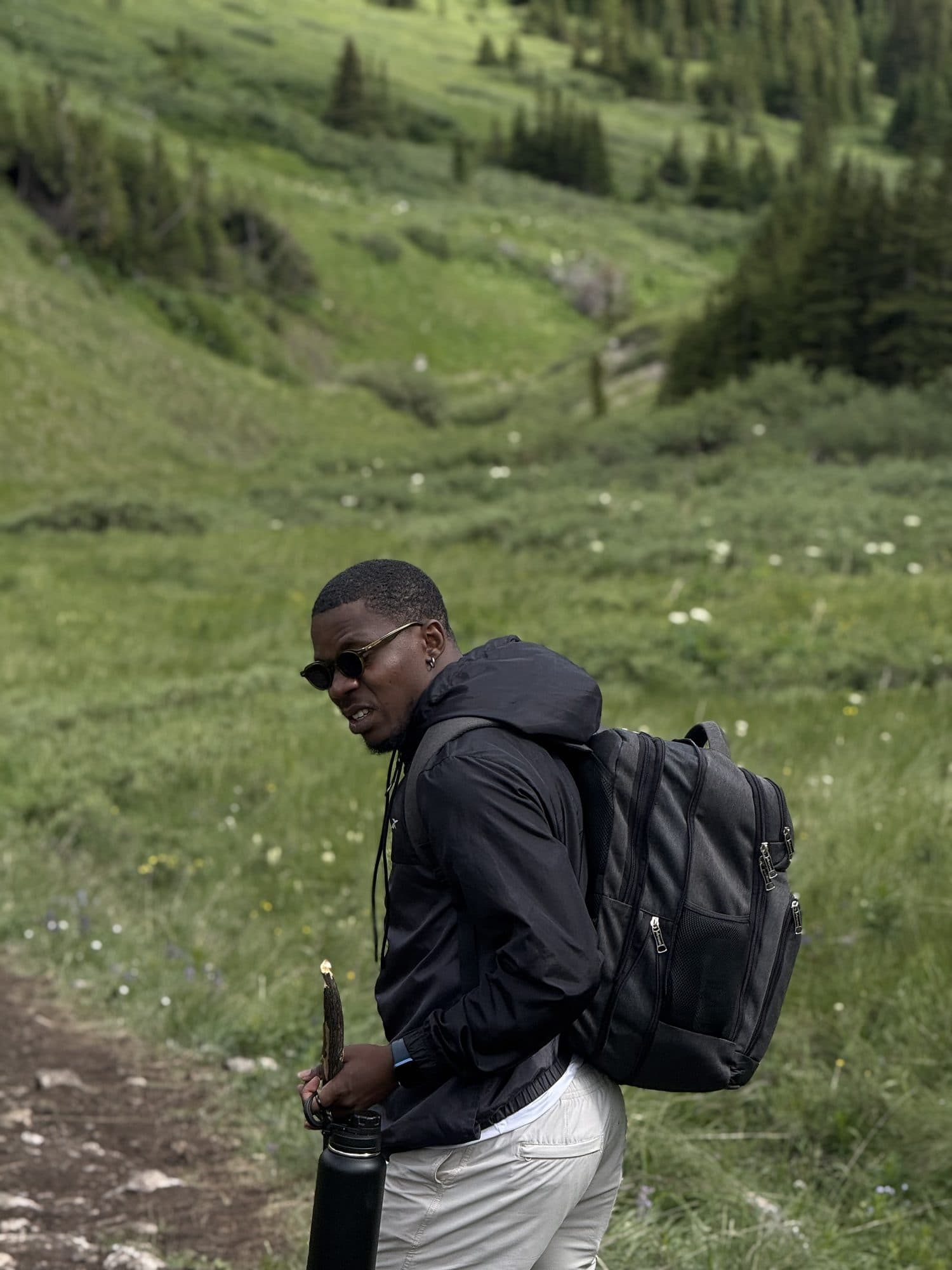 Femi on a green mountain trail with a backpack and sunglasses, glancing back
