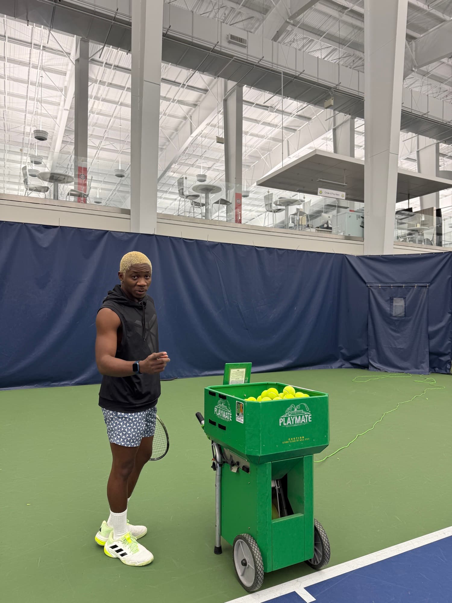 Standing beside a green Playmate ball machine on an indoor court